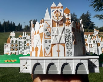 Könnte beinhalten: Ein detailliertes 3D-gedrucktes Modell eines skurrilen Gebäudes mit weißen, grauen und orangefarbenen Akzenten. Das Modell weist komplizierte architektonische Details auf, darunter Bögen, Türme und dekorative Elemente. Das Gebäude befindet sich vor dem Hintergrund von grünem Gras und blauem Himmel.