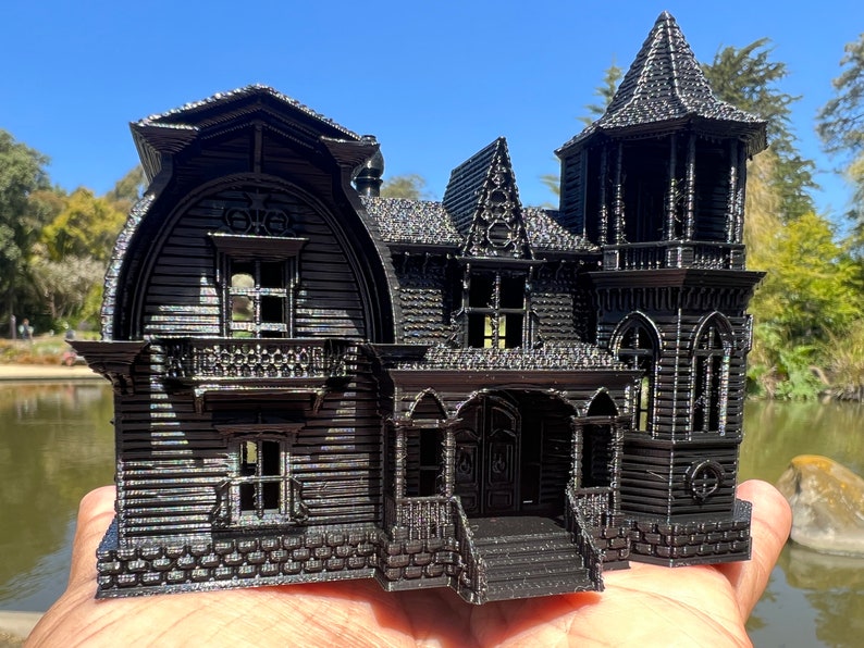 May include: A black 3D printed model of a Victorian style house with a tower, a porch, and a stone foundation.