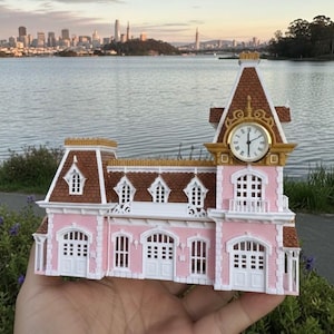 Puede incluir: Un edificio en miniatura rosa y blanco con una torre del reloj. El edificio tiene un tejado marrón, adornos blancos y una esfera dorada. El reloj marca la 1:00. El fondo presenta una masa de agua y un horizonte urbano.