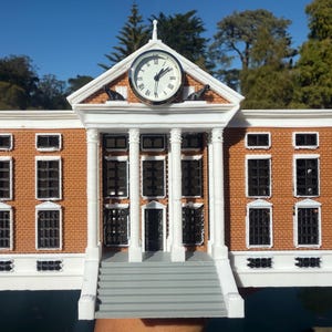 May include: A miniature model of a building with a clock tower. The building has a brick facade, white columns, and a clock with black hands. The model is held up against a background of trees and a blue sky.