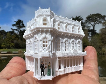 Könnte beinhalten: Ein detailliertes weißes Miniaturhaus mit aufwendigen architektonischen Details, darunter Fenster und ein dekoratives Dach. Zwei Miniaturfiguren stehen im Türrahmen. Das Haus wird in einer Hand gehalten, mit einem Park und einem See im Hintergrund.