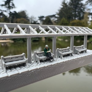 May include: A miniature gray 3D printed model of a park gazebo with a trellis roof and three benches. A small figurine of a person is sitting on one of the benches.