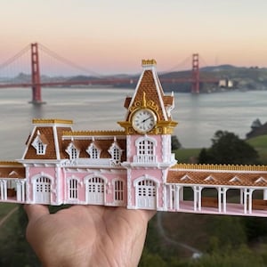 Puede incluir: Un edificio en miniatura rosa y blanco con una torre de reloj, techo marrón y detalles dorados. El modelo se sostiene frente al puente Golden Gate y la bahía de San Francisco. El edificio tiene múltiples ventanas y una pasarela cubierta.