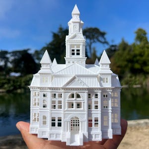 Könnte beinhalten: Ein weißes 3D-gedrucktes Modell eines Gebäudes mit einem Uhrturm und filigranen Details. Das Modell wird in einer Hand vor einem verschwommenen Hintergrund aus einem Teich und Bäumen gehalten.