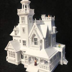 May include: A white 3D printed model of a Victorian style house with a tower, porch, and several windows. There are small figures of people standing on the roof and porch.