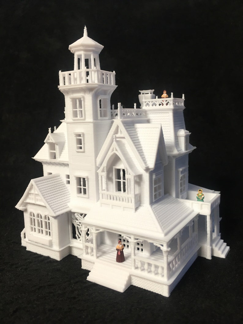 May include: A white 3D printed model of a Victorian style house with a tower, porch, and several windows. There are small figures of people standing on the roof and porch.