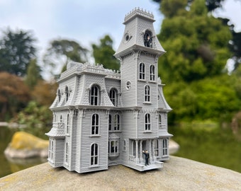 May include: A gray miniature Victorian house with multiple stories, windows, and a tower. A small figure stands on the porch, and another is visible in the tower. The house is set against a blurred background of trees and water.