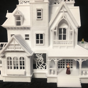 May include: White 3D printed model of a Victorian house with a porch, a greenhouse, and two miniature figures. The house has a detailed facade with windows, a roof, and decorative trim.