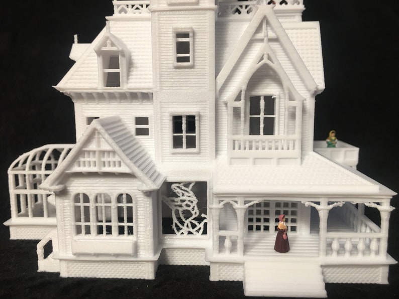May include: White 3D printed model of a Victorian house with a porch, a greenhouse, and two miniature figures. The house has a detailed facade with windows, a roof, and decorative trim.