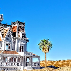 May include: A white Victorian-style house with a red roof and a tower, surrounded by desert landscaping and a white gazebo with a red roof.
