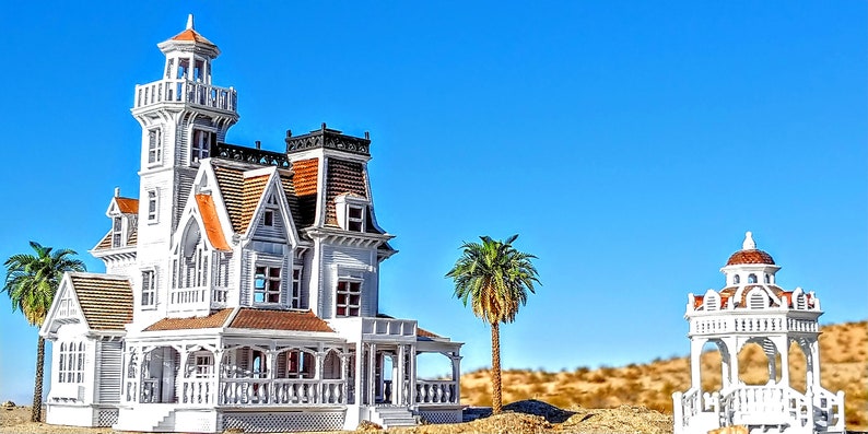 May include: A white Victorian-style house with a red roof and a tower, surrounded by desert landscaping and a white gazebo with a red roof.