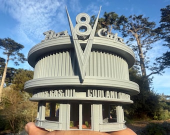 May include: A gray, 3D-printed model of a retro-style diner, featuring the text "Flat 8 Cafe" and "Fuels Gas Oil Coolant Grease." The design includes a prominent "V" shape and the number "8" in the center, with a rounded, art deco aesthetic.