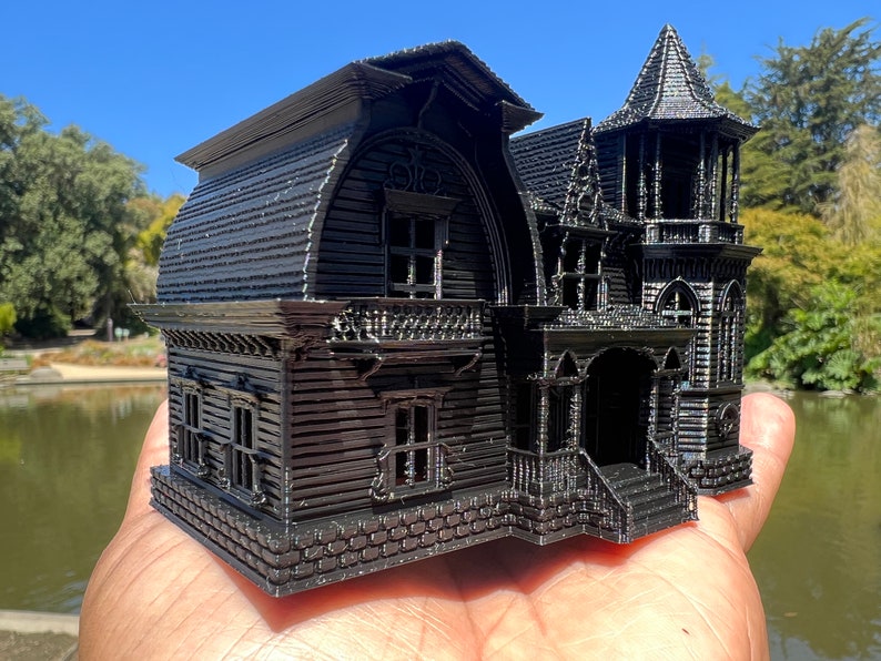 May include: A black 3D printed model of a Victorian style house with a steep roof, multiple windows, and a front porch. The model is detailed with intricate trim and brickwork.