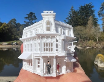 May include: White 3D printed model of a Victorian-style building with a sign that reads "Baker". The model is detailed with windows, doors, and a balcony. Two miniature figures stand in front of the building.