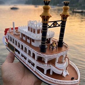 May include: A detailed model of a paddle steamer boat, featuring white, brown, and black accents. The boat has two tall smokestacks with gold tops, a red paddle wheel, and multiple decks with intricate railings. The model is held against a blurred water background.