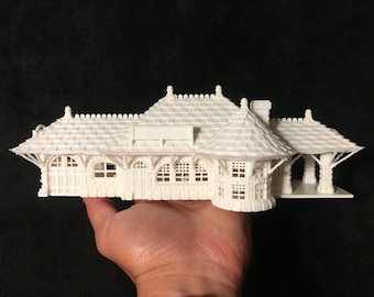 May include: A detailed white model of a train station, featuring intricate architectural details such as arched windows, a tiled roof, and decorative trim. The model is held against a dark background, showcasing its design.