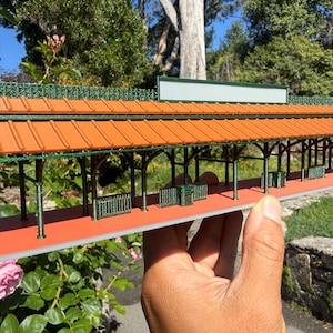 May include: A miniature model of a train station with an orange roof, green trim, and a red platform. The model has detailed architectural features, including support columns and benches. The station is held in front of a natural background of greenery and a blue sky.