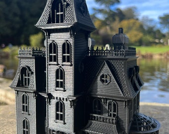 May include: A black 3D printed model of a Victorian-style house with a gabled roof, multiple windows, and a small greenhouse addition. The house is detailed with intricate trim and a small figure standing on the front porch.