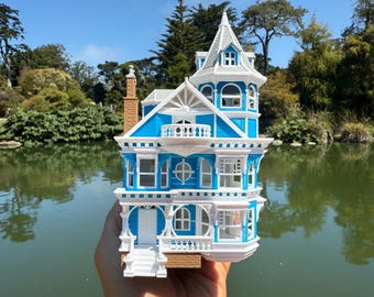 May include: A detailed miniature Victorian house with a blue and white color scheme. The model features intricate architectural details, including a tower, balconies, and decorative trim. The house is held in front of a lake with trees in the background.