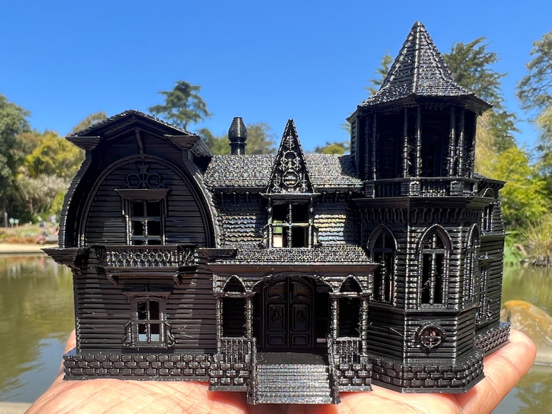 May include: A black 3D printed model of a gothic-style house with a tower, multiple windows, and a front porch. The house is detailed with intricate trim and a brick foundation.