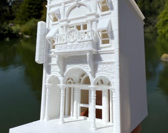 May include: A white, 3D-printed model of a building with architectural details, including arched entryways, columns, and a balcony with the word "Arcade". The model has a detailed facade and is held against a backdrop of water and trees.