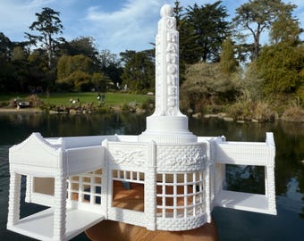 May include: A white 3D-printed model of a monument with the text "RAMONES" vertically displayed. The model features a detailed architectural design, including a base, columns, and a decorative top. The background shows a lake and trees.