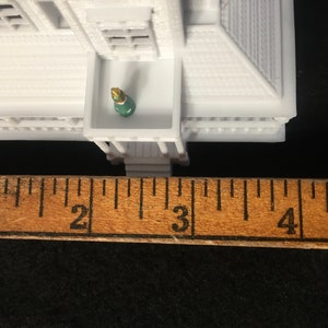 May include: White miniature house with a green and gold figure standing on a balcony. The house is measured with a ruler, showing it is approximately 2.5 inches tall.