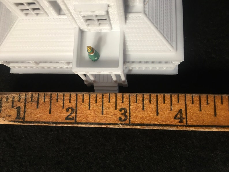 May include: White miniature house with a green and gold figure standing on a balcony. The house is measured with a ruler, showing it is approximately 2.5 inches tall.