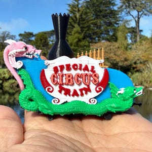Op de afbeelding: Een kleurrijk 3D-geprint circus treinspeelgoed. De trein heeft een blauwe basis, groene drakenfiguren en een roze drakenkop. Het bord leest "SPECIAL CIRCUS TRAIN" in rode letters. Een zwarte toren staat erachter.