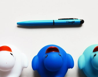 Può includere: Tre anatre di gomma, una bianca, una blu e una azzurra, con becchi rossi e creste arancioni. Le anatre sono disposte in fila su una superficie bianca. Una penna blu con uno stilo nero è sopra le anatre.