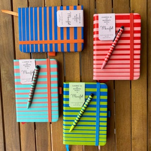 Può includere: Quattro quaderni a righe colorati con penne abbinate. Ogni quaderno ha un'etichetta che dice "CUADERNO AS con BOLÍGRAFO metálico by Manifol" e "PLAN ACID FREE PAPER 90 G/M2 SIZE ONE FEEL".