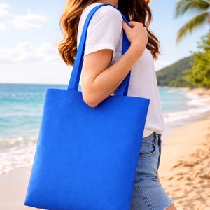 May include: A vibrant blue tote bag with matching straps is held by a person on a beach. The bag is rectangular and appears to be made of a sturdy fabric. The background shows a sandy beach, ocean, and blue sky.