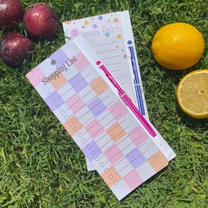 Könnte beinhalten: Zwei rosa-weiße karierte Einkaufslisten mit dem aufgedruckten Text "Shopping List". Eine Liste ist leer, die andere hat ein paar darauf geschriebene Punkte. Auf den Listen liegen zwei Stifte, einer blau und einer rosa. Die Listen liegen auf einem Bett aus grünem Gras.