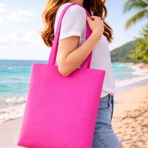 May include: A bright pink tote bag with matching straps is held over the shoulder. The bag is rectangular and appears to be made of a sturdy fabric. The background shows a beach scene with blue water and a sunny sky.