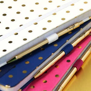 Puede incluir: Una pila de cuadernos en blanco, azul marino y rosa, cada uno con lunares dorados. Bolígrafos dorados con la palabra "Manifest" están sujetos a los cuadernos. Los cuadernos tienen bandas elásticas y bordes dorados.