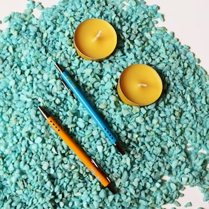 May include: Two yellow beeswax candles and two pens, one blue and one orange, on a bed of turquoise blue pebbles.