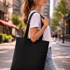 May include: A solid black tote bag with long handles, carried over the shoulder. The bag is rectangular and appears to be made of canvas or a similar fabric. The background shows a city street with buildings and trees.