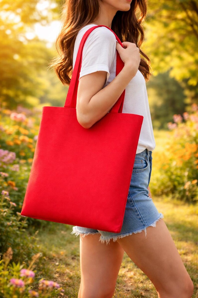 May include: A solid red tote bag with matching red handles is held by a person. The bag is rectangular and appears to be made of a sturdy fabric. The person is wearing a white t-shirt and denim shorts.