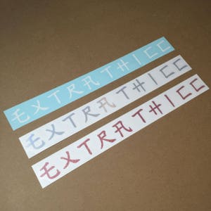 May include: Three vinyl stickers with the text "EXTRATHICC" in different colours and fonts. The top sticker is light blue, the middle sticker is light grey, and the bottom sticker is pink.
