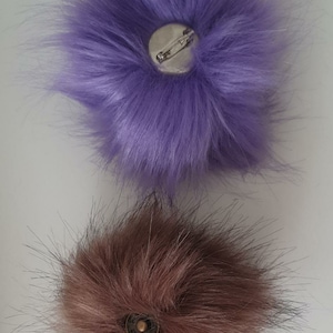 May include: Two fluffy pom poms with metal clasps on the back. The top pom pom is purple and the bottom pom pom is brown.