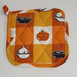 Op de afbeelding: Oranje en witte gewatteerde ovenwant met een geruit patroon en zwart-witte illustraties van verschillende keukengerei, waaronder een tomaat, een pan en een aubergine. De ovenwant heeft een lus om op te hangen en een Le Creuset-label.