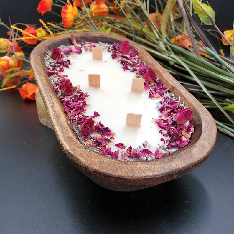 May include: A large, wooden dough bowl candle filled with white wax and four wooden wicks. The candle is decorated with dried rose petals and herbs. The bowl is a warm brown color, and the background is dark.