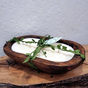 May include: A white candle in a wooden dough bowl with a green leafy garland wrapped around it. The candle has three wicks and is surrounded by the garland. The bowl is brown and has a rustic look.