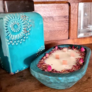 May include: A teal-colored, wooden-style candle holder with a creamy candle, surrounded by dried rose petals and pink buds. The candle has four wooden wicks. A teal ceramic jar with a decorative pattern is in the background.