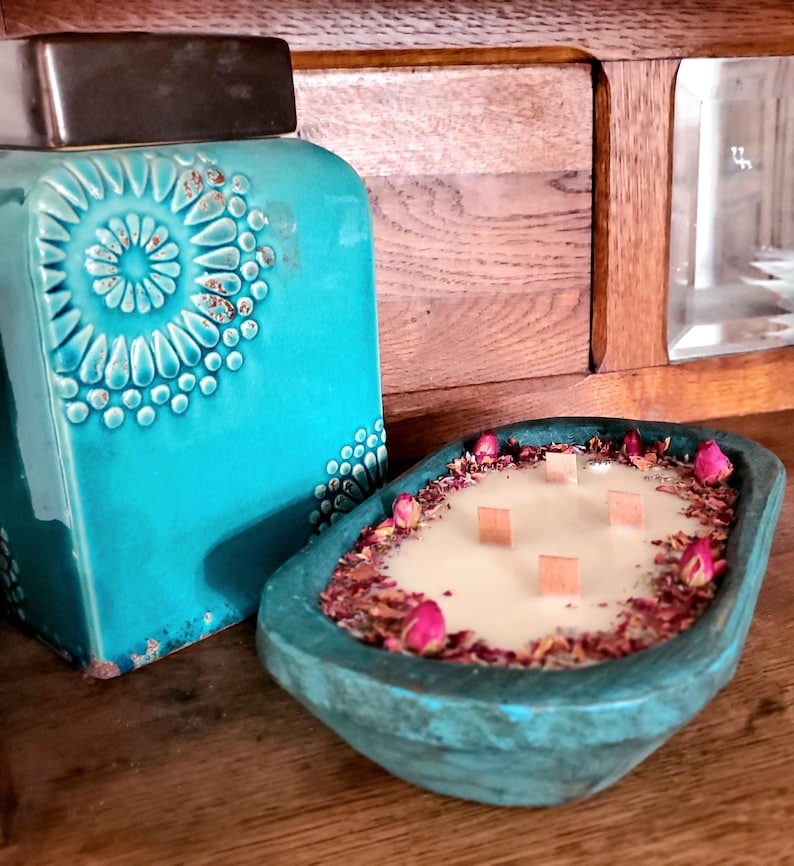 May include: A teal-colored, wooden-style candle holder with a creamy candle, surrounded by dried rose petals and pink buds. The candle has four wooden wicks. A teal ceramic jar with a decorative pattern is in the background.