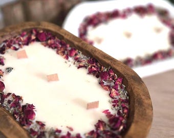 May include: A wooden dough bowl candle filled with white wax, adorned with dried rose petals and wooden wicks. The candle has a rustic aesthetic, with a second candle in the background. The candle is a decorative home fragrance item.