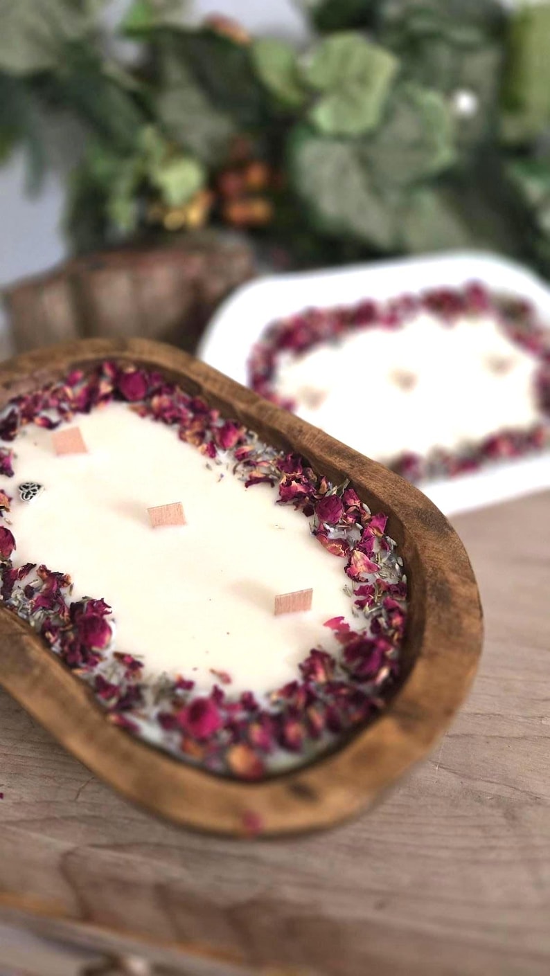 May include: A wooden dough bowl candle filled with white wax, adorned with dried rose petals and wooden wicks. The candle has a rustic aesthetic, with a second candle in the background. The candle is a decorative home fragrance item.