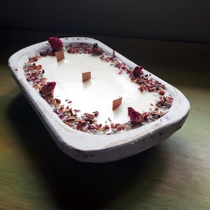 May include: A white, rectangular candle in a rustic wooden bowl. The candle has four wooden wicks and is decorated with dried rose buds and petals. The bowl sits on a green surface.