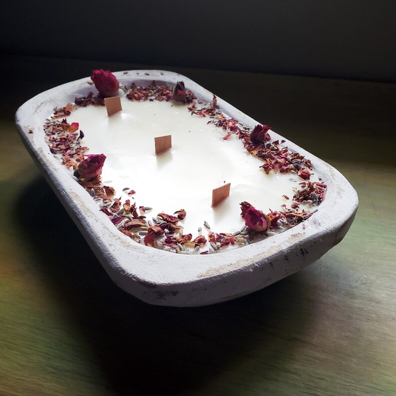 May include: A white, rectangular candle in a rustic wooden bowl. The candle has four wooden wicks and is decorated with dried rose buds and petals. The bowl sits on a green surface.