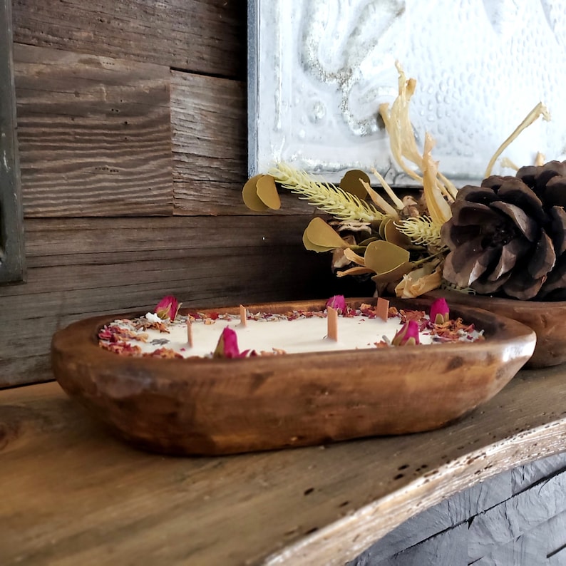 May include: A wooden dough bowl candle with a creamy white wax, adorned with dried rose petals and buds. The candle has four wooden wicks and is placed on a wooden surface. The bowl is a warm brown color.